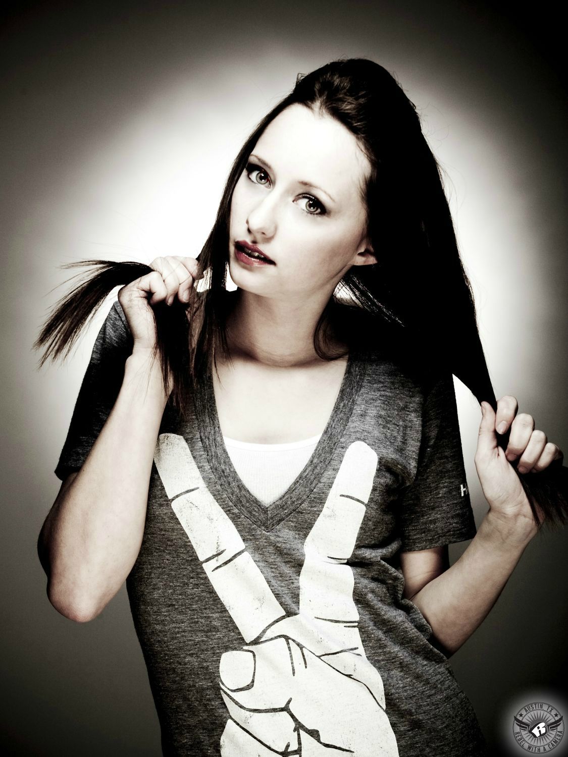 brunette female fashion model with fair skin, green eyes and red lipstick with a grey low cut shirt with a hand image on it giving the peace sign pulls own hair in a photo studio in austin with a white background